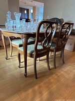 Photo showing oval wooden dining table with curved legs and scalloped edges, surrounded by four chairs. Crystal glassware on tabletop (not included).
