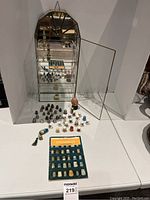 Glass and metal display case open showing collection of thimbles, booklet, cloisonné elephant, and small decorative items arranged on white surface.