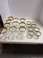 Overall view of 18 glasses grouped on white surface showing gold rims and etched patterns; 10 wine glasses and 8 cocktail glasses.