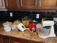 Countertop with kitchen appliances and cookware. Philips crock pot, Sunbeam rice cooker, Mixmaster mixer, hand mixer, Corning Ware dishes visible.