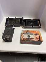 Three vintage cameras including two Kodak folding pocket cameras and one Keystone instant loading color camera in original box displayed on a white surface.
