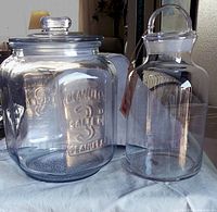 Two clear glass apothecary style jars on cloth surface, one embossed with text, other plain with handle lid.