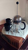 Full view of Bodum French press with two black ceramic mugs on a cloth on a wooden surface.