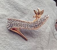 Front angle photo of sparkly high-heel shoe brooch showing rhinestone detail and bow on gold tone metal.