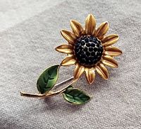 Front view showing gold-tone sunflower petals, chocolate brown rhinestone center, and green enamel leaves on stem.