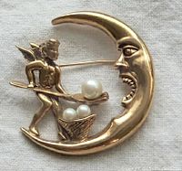 Close-up image of the gold-tone metal artistic brooch depicting a crescent moon with a face and an angel feeding faux pearls.