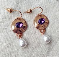 Pair of gold-toned hammered metal drop earrings with oval faceted purple amethyst stones and teardrop white pearls shown on fabric background.