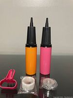 Two balloon pumps in orange and pink with black nozzles, placed on a reflective surface with white tapes and a pink clip in front.