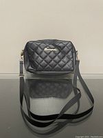 Black quilted handbag purse on a reflective surface, showing front view with brand logo and shoulder strap.