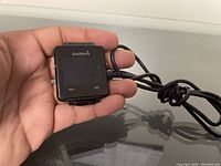 Close-up photo of Garmin smartwatch held in hand with charger cable attached, showing screen and Garmin branding.