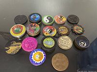 Photo of 17 metal pogs with various designs including yin yang, Bigfoot, 8-ball, official stamps, and cartoon images, displayed on black surface.
