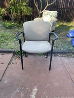 Front angled view of grey fabric office chair with black frame and armrests outdoors on a paved area with grass and plants in the background.