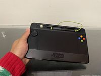 Tablet held in hand showing front with directional pad, colored buttons and stylus at top.