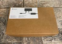 Photo of the original brown box packaging with a white label showing the Xbox Kinect adapter power supply diagram and multilingual text.