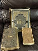 Three antique books arranged on a dark leather couch, showing the covers with the largest book titled 'The Holy Bible', a second book titled 'Byron', and a smaller worn book without visible title on the cover.