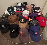 Photo showing 20 various ball caps including sports team logos and novelty designs arranged on a round table.