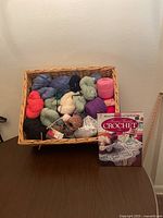 Wicker basket filled with various skeins of yarn and a crochet book placed beside it.