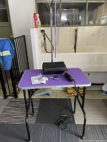 Full view of purple dog grooming table with Oster A5 clippers and blades on top, folding black legs visible, adjustable grooming arm attached.