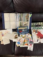 Photo of a large binder with postcards in protective sleeves, loose postcards and vintage greeting cards, and advertisement cards laid out on a couch.