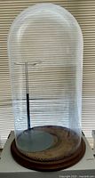 Clear large glass dome on a wooden base with metal doll stand and antique braided mat inside, photographed against window blinds.