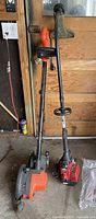 Two gardening tools: an electric lawn edger with orange housing and wheels, and a red gas-powered weed whacker with curved shaft and black handle, leaned against a wooden door.