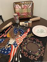 Overview of the lot items including the souvenir wall hanging, jar of dice, loom kit components, throw blanket in carrier, and memorial plate on a table.