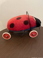 Side view showing red and black plush ladybug body with black spots on metal frame and four wheels.