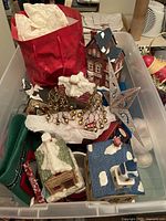 Plastic tub containing vintage Christmas decorations such as ceramic houses with snow accents, glittery star, and metallic garlands.