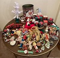 Overview of entire lot showing all Christmas themed items displayed on a round wooden table, including a Santa-themed tin, white fabric tree, various fabric and ceramic ornaments, and Dept 56 boxed items.