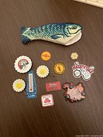 Top-down image showing the fish skillet handle cover and all ten vintage refrigerator magnets arranged on a wooden surface with a ruler for scale.