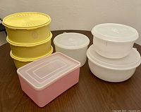Photo of seven vintage Tupperware containers in yellow, white, and pink colors arranged on a wooden surface.