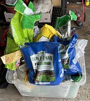 Plastic tub filled with multiple partially used bags of lawn seed mixes, weed and feed, and potting soil in various colors such as blue, yellow, green, and white.