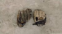 Photo of two baseball gloves placed on a light gray textured surface. One glove is dark brown and the other is light beige with black details.
