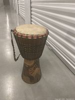 Full view of the wooden hand-carved djembe drum standing on concrete floor with a focus on the carved patterns and drumhead.