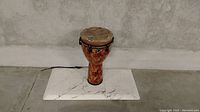 Single photo showing a djembe style bongo drum with red decorative pattern and natural skin head, placed on a white marble slab against a concrete wall