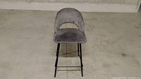 Front view of gray velvet bar stool chair with black metal legs and footrest, placed on concrete floor.