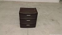 Brown plastic 3-drawer storage unit shown from the front on a concrete floor.
