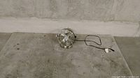 Small silver metal desk fan with three blades and protective wire cage, power cord and plug attached, placed on concrete surface.