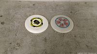 Photo of two white frisbees on grey surface, showing their colorful center designs clearly.