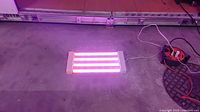 Grow light fixture emitting pinkish light with 4 fluorescent tubes on concrete floor, connected to power cord and extension cable.