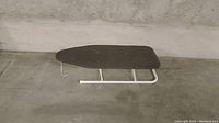 Side view of the ironing board showing padded surface and metal legs folded down on concrete floor.