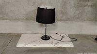 Black lampshade table lamp with glass and metal stand on a marble slab against concrete wall