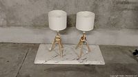 Two small wooden tripod table lamps with white drum shades on a marbled rectangular slab against concrete wall.