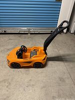 Side view of the orange McLaren push car with black push handle attached.