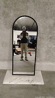 Mirror with rounded arch top and black beaded frame, shown upright against a concrete wall, reflected image shows person taking photo.