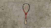 Front view of the Prince red tennis racket laid on a sandy surface, showing the entire racket including the head, strings, throat, and grip wrapped in worn beige tape.