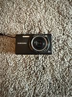 Black Samsung compact digital camera placed on beige carpet showing front with lens and built-in flash.