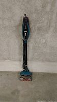 Photo of a blue and black Dirt Devil Duo Clean upright vacuum cleaner with power cord neatly wrapped, shown standing upright against a concrete wall and floor.