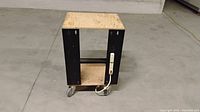 Photo of a mobile work bench with plywood top, black metal frame, bottom shelf, caster wheels, and power strip attached to one side.
