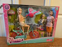 Full boxed set showing the three dolls, teal bicycle with red wheels, and ball accessory in the packaging with branding and age recommendation.